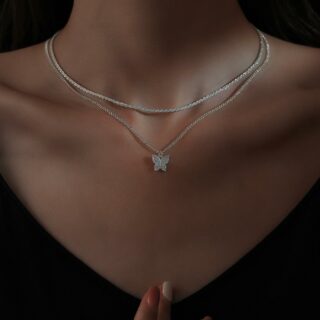 pendentif en forme de papillon argenté et délicat, à double chaîne argentée, le tout porté par une silhouette de femme avec col et cou visible, portant un habit noir, dans une lumière sombre, sur fond noir