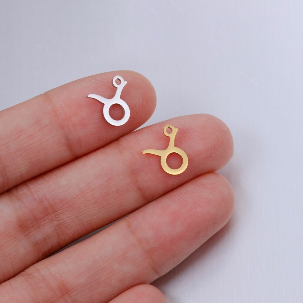 deux pendentifs en forme de signe du taureau, un argenté un doré, sur le bout de deux doigts d'une main blanche, sur fond gris