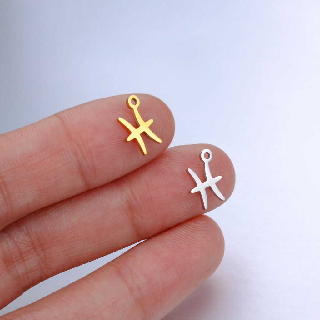 deux pendentifs en forme de signe des poissons, un argenté un doré, sur le bout de deux doigts d'une main blanche, sur fond gris