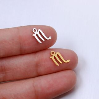 deux pendentifs en forme de signe du scorpion, un argenté un doré, sur le bout de deux doigts d'une main blanche, sur fond gris