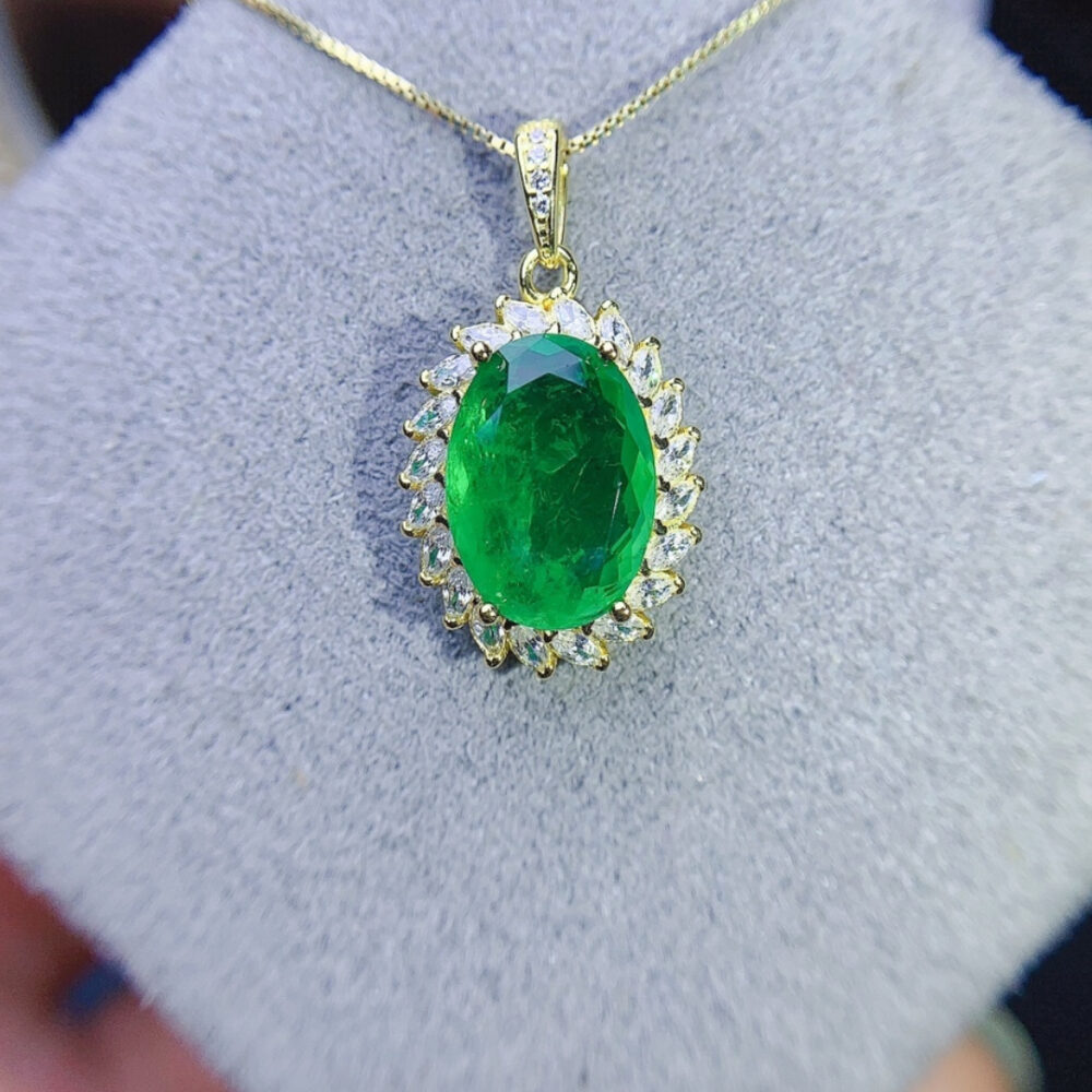 Pendentif ovale avec une émeraude verte au centre, fond gris.