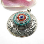 pendentif tibétain rond, en métal argenté gravé de détails de fleurs et de spirales, avec pierres en différents cercles, bleu/vertes, rouges et une bleu foncée au centre, sur fond blanc