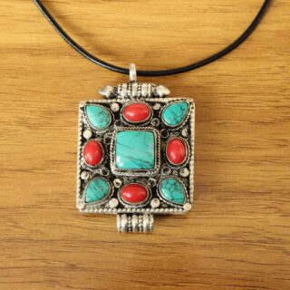 pendentif carré tibétain en argent avec pierres semi précieuses placées de façon géométrique, une grosse carrée centrale cleu clair, 4 plus petites bleu clair dans les coins du carré, et 4 rouges sur les côtés. Il est attaché à un fil de collier noir, sur fond de parquet marron clair.