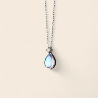 Collier en argent avec chaîne fine et pendentif goutte en pierre de lune irisée, monté sous un zircon brillant ; sur fond beige.