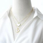 2 colliers dorés avec pendentif cœur en nacre blanche sur un buste en tissu gris clair qui porte une chemise ouverte blanche.