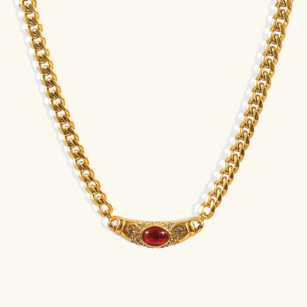 Collier doré avec pendentif ovale vintage, pierre rouge brillante au centre, chaîne à mailles épaisses ; sur fond neutre.