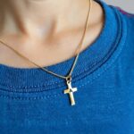 Pendentif en forme de croix dorée avec initiale gravée porté par un enfant qui porte un t-shirt bleu