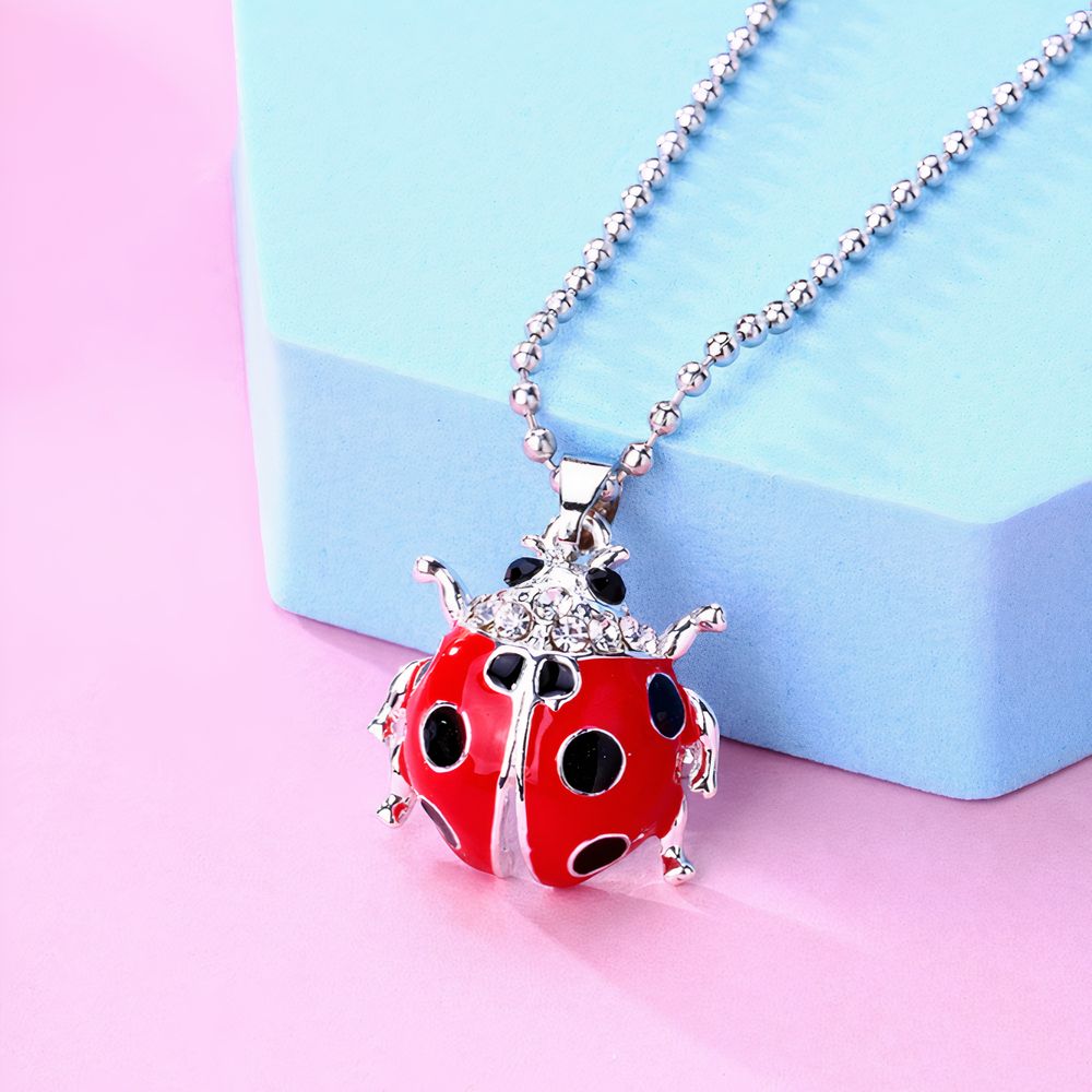 Collier enfant avec pendentif coccinelle rouge et argent en émail, sur une chaîne à billes argentée. Le fond est un objet bleu simple posé sur un sol rose.