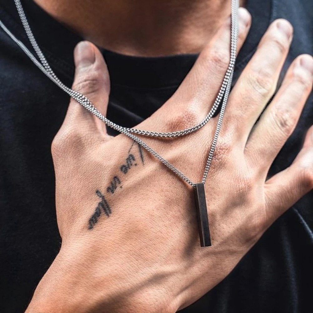 Collier homme avec pendentif rectangulaire brillant suspendu à une chaîne fine, posé sur la main d'un homme qui porte la chaine autour du cou.