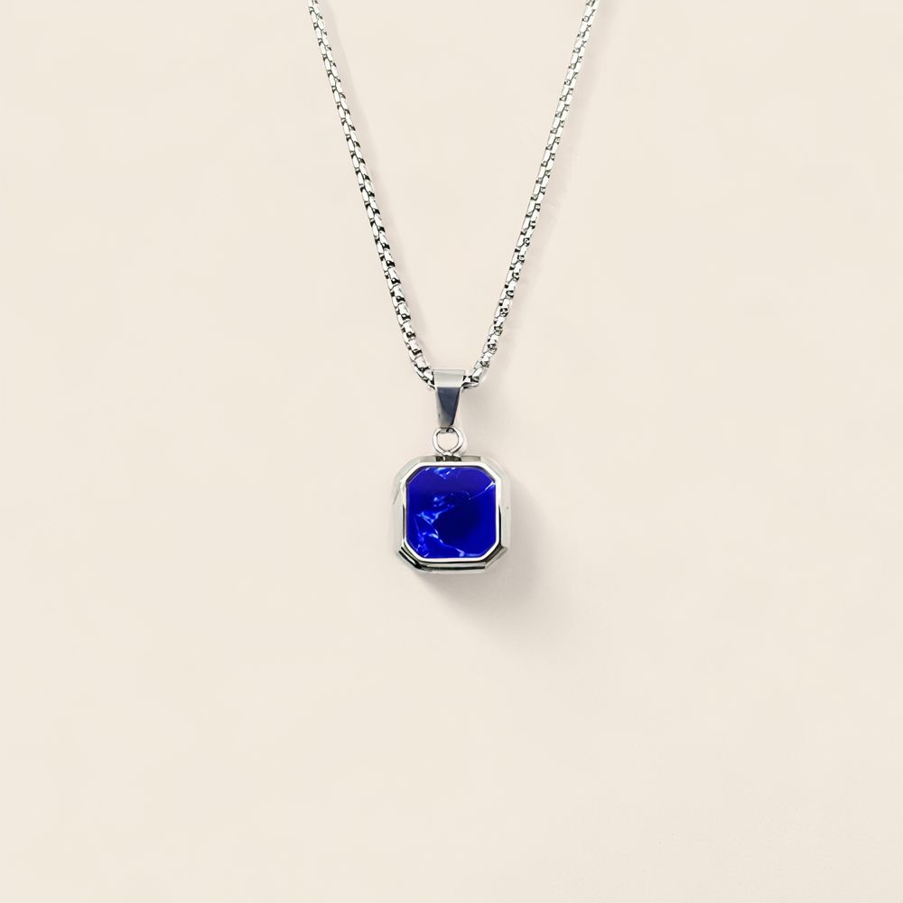 Pendentif homme en acier couleur argent avec pierre carrée colorée bleu foncé, sur chaîne épaisse argentée ; sur fond beige.