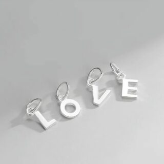 Petits pendentifs en forme de lettres majuscules avec anneaux, en argent 925 brillant, posés sur fond gris.
