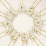 pendentifs doré en forme de lettres arrondie incrustée de strass, suspendu à une chaîne fine brillante en acier inoxydable, en cercles , sur fond beige.