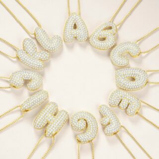 pendentifs doré en forme de lettres arrondie incrustée de strass, suspendu à une chaîne fine brillante en acier inoxydable, en cercles , sur fond beige.