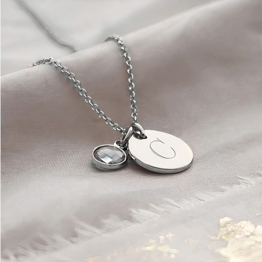 Pendentif rond argenté gravé d’une lettre, avec petite pierre colorée en pendentif assorti , sur une chaîne argentée ; le tout est posé sur un tissu gris.