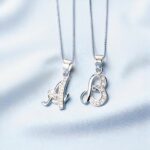 pendentifs lettres en argent en majuscule cursive, ornés de petits zircons, disposés sur fond bleu clair