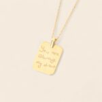 pendentif en forme de plaque dorée avec message manuscrit gravé, chaîne fine couleur or ; sur fond beige.
