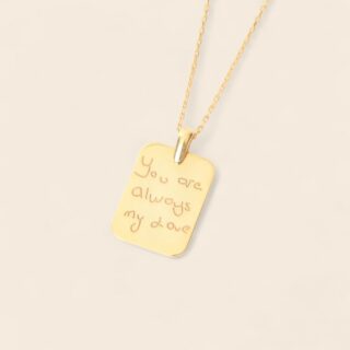 pendentif en forme de plaque dorée avec message manuscrit gravé, chaîne fine couleur or ; sur fond beige.