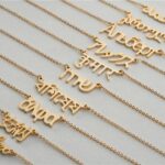pendentifs personnalisés avec prénoms en différentes langues (arabe, hindi, coréen…), sur chaînes fines dorées, sur fond gris.